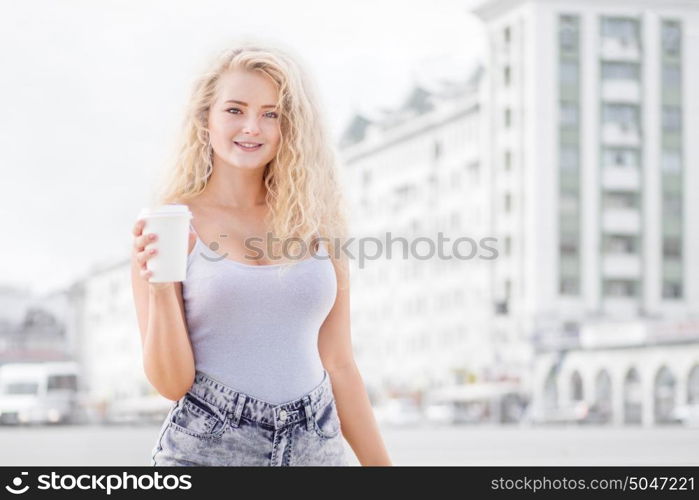 Happy young woman with long curly hair, holding a take away coffee cup and smiling with flirt in front of a camera against urban city traffic background.