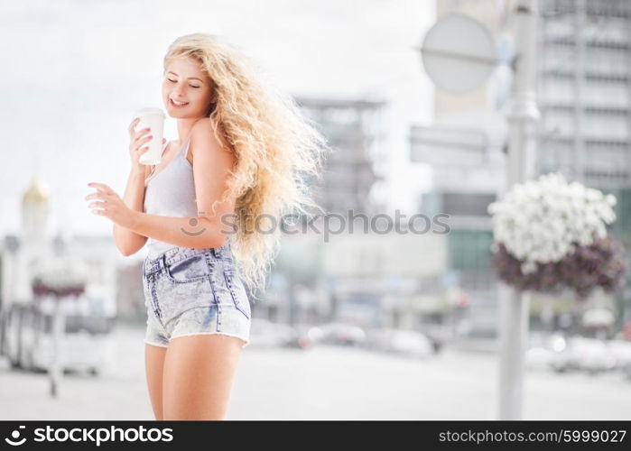 Happy young woman with long curly hair, holding a take away coffee cup and smiling with flirt in front of a camera against urban city traffic background.