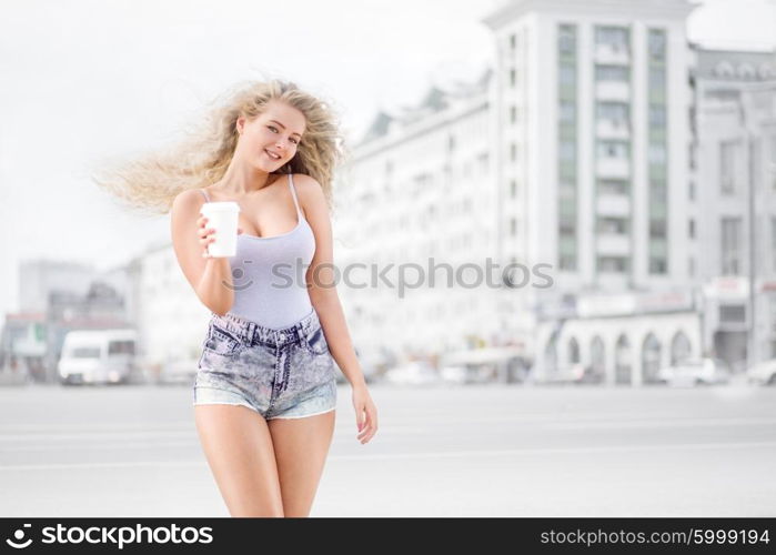 Happy young woman with long curly hair, holding a take away coffee cup and smiling with flirt in front of a camera against urban city traffic background.