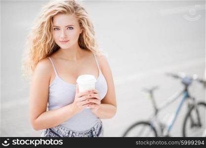 Happy young woman with long curly hair, holding a take away coffee cup and standing against empty road and city bike background.