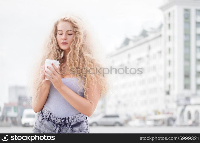 Happy young woman with long curly hair, holding a take away coffee cup and smiling with flirt in front of a camera against urban city traffic background.