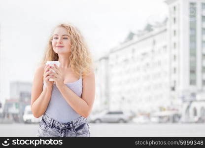 Happy young woman with long curly hair, holding a take away coffee cup and smiling with flirt in front of a camera against urban city traffic background.