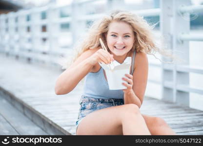 Happy young woman with long curly hair, sitting on the wooden floor, holding a lunch box and eating up noodles from Chinese take-out with chopsticks.