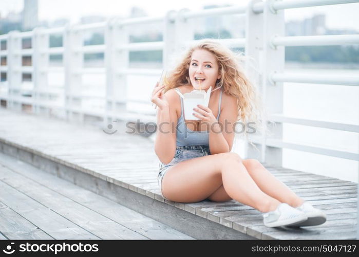 Happy young woman with long curly hair, sitting on the wooden floor, holding a lunch box and eating up noodles from Chinese take-out with chopsticks.