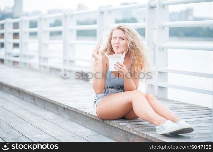 Happy young woman with long curly hair, sitting on the wooden floor, holding a lunch box and eating up noodles from Chinese take-out with chopsticks.