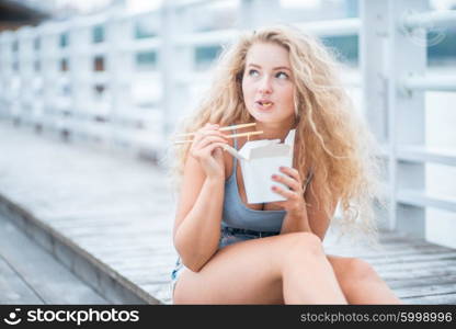 Happy young woman with long curly hair, sitting on the wooden floor, holding a lunch box and eating up noodles from Chinese take-out with chopsticks.