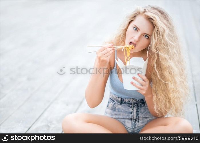 Happy young woman with long curly hair, sitting on the wooden floor, holding a lunch box and eating up noodles from Chinese take-out with chopsticks.
