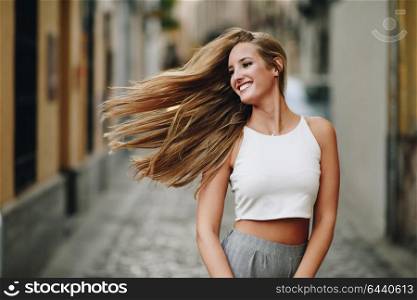 Happy young woman with moving hair in urban background. Blond girl, straight hairstyle, wearing casual clothes in the street.
