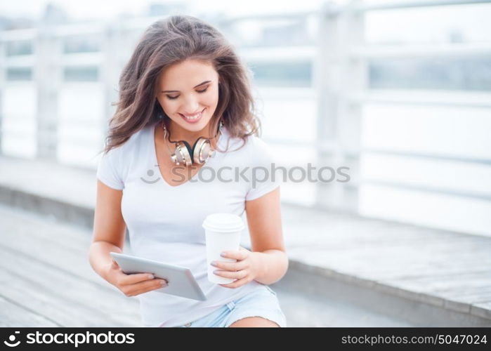 Happy young woman with music headphones and a take away coffee cup, surfing internet on tablet pc, listening to the music and sitting on the bridge.