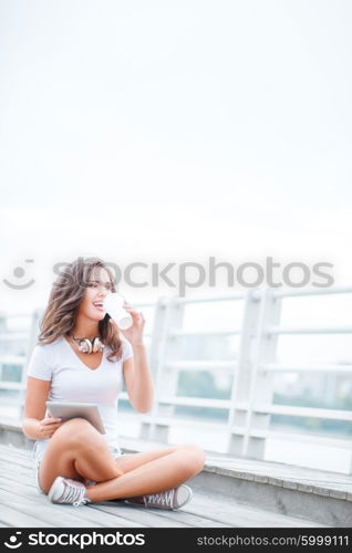 Happy young woman with music headphones and a take away coffee cup, surfing internet on tablet pc, listening to the music and sitting on the bridge.