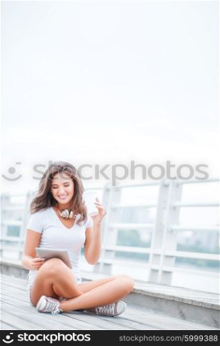 Happy young woman with music headphones and a take away coffee cup, surfing internet on tablet pc, listening to the music and sitting on the bridge.