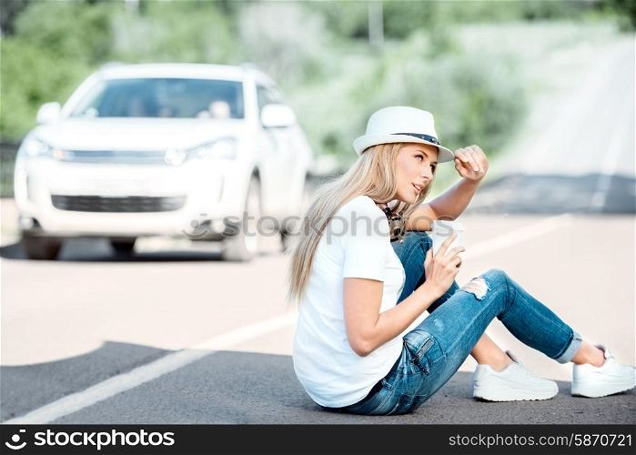 Happy young woman with music headphones around her neck, drinking coffee from a takeaway coffee cup and sitting on a separating strip against road background.