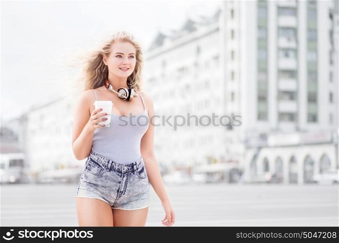 Happy young woman with music headphones, holding a take away coffee cup, listening to the music and walking against city background.