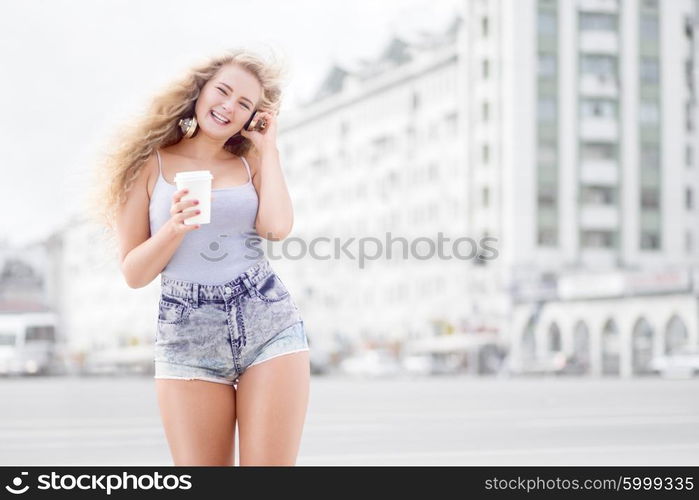 Happy young woman with music headphones, holding a take away coffee cup, listening to the music and walking against city background.