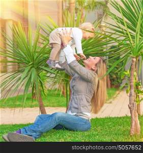 Happy young woman with pleasure playing outdoors with her cute little blond daughter, mother lifting up baby girl, having fun in the garden in sunny day, love and enjoyment concept