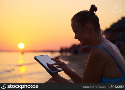 Happy young woman with tablet PC on beach at sunset. Evening sun, sea and beach on background