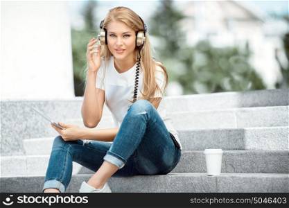 Happy young woman with vintage music headphones and a take away coffee cup, surfing internet on tablet pc, listening to the music and sitting on stairs against urban city background.