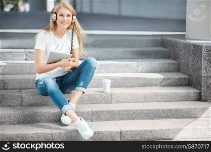 Happy young woman with vintage music headphones and a take away coffee cup, surfing internet on tablet pc, listening to the music and sitting on stairs against urban city background.