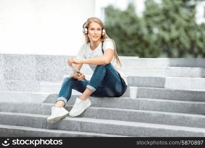 Happy young woman with vintage music headphones and a take away coffee cup, surfing internet on tablet pc, listening to the music and sitting on stairs against urban city background.