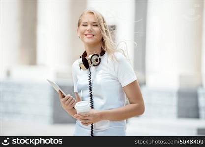 Happy young woman with vintage music headphones around her neck, holding a take away coffee cup with a straw and a tablet pc, standing against urban city.