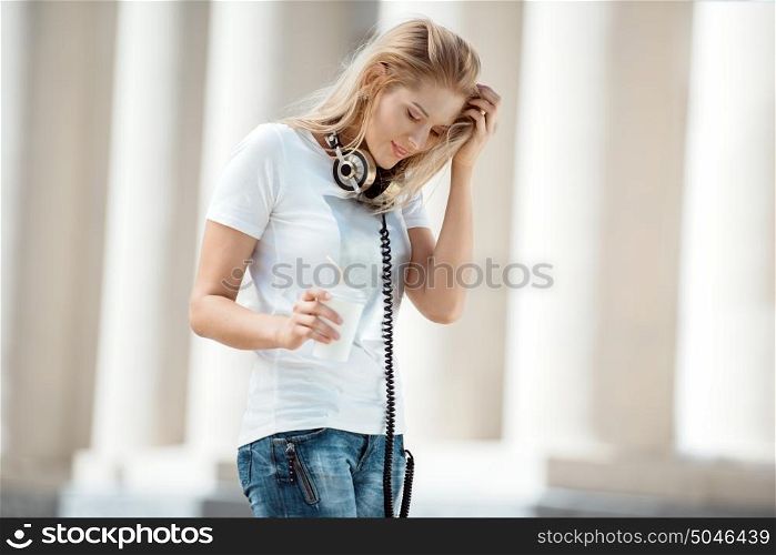 Happy young woman with vintage music headphones around her neck, holding a take away coffee cup with a straw and a tablet pc, standing against urban city.
