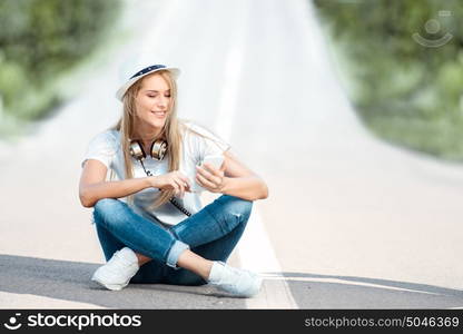 Happy young woman with vintage music headphones around her neck, surfing internet on a smartphone and sitting on a separating strip against road background.