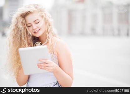 Happy young woman with vintage music headphones around her neck, surfing internet on a tablet pc and posing against urban city background.