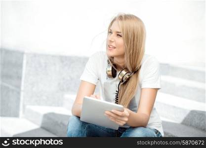 Happy young woman with vintage music headphones around her neck, surfing internet on a tablet pc and sitting on stairs against urban city background.