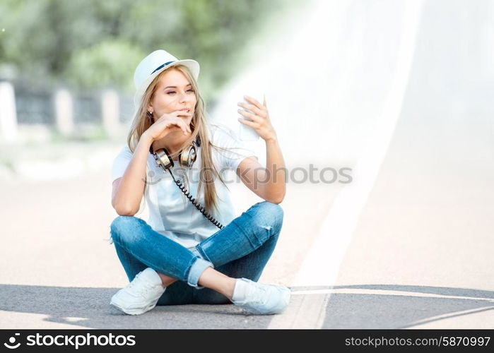 Happy young woman with vintage music headphones around her neck, surfing internet on a smartphone and sitting on a separating strip against road background.