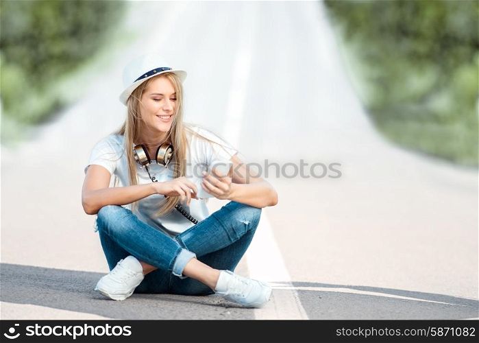 Happy young woman with vintage music headphones around her neck, surfing internet on a smartphone and sitting on a separating strip against road background.