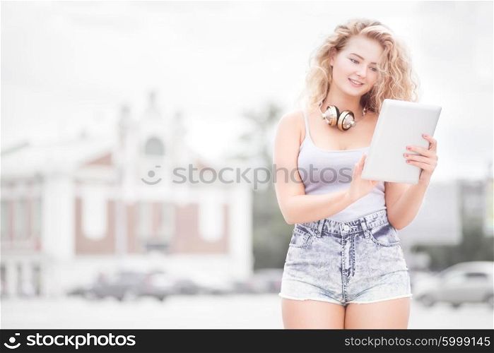 Happy young woman with vintage music headphones around her neck, surfing internet on a tablet pc and posing against urban city background.