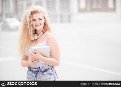 Happy young woman with vintage music headphones around her neck, surfing internet on a tablet pc and posing against urban city background.