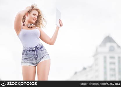 Happy young woman with vintage music headphones around her neck, taking selfie with tablet pc and smiling happily against urban city background.