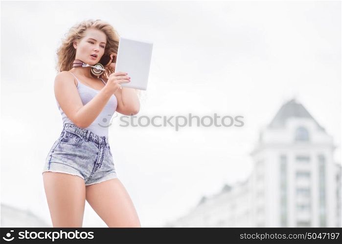 Happy young woman with vintage music headphones around her neck, taking selfie with tablet pc and smiling happily against urban city background.