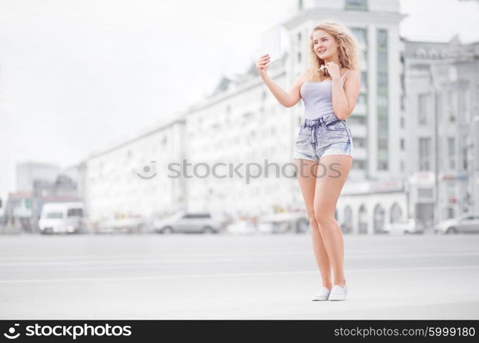 Happy young woman with vintage music headphones around her neck, taking selfie with tablet pc and smiling happily against urban city background.