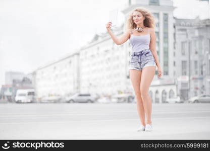 Happy young woman with vintage music headphones around her neck, taking selfie with tablet pc and smiling happily against urban city background.