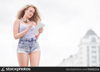 Happy young woman with vintage music headphones around her neck, taking selfie with tablet pc and smiling happily against urban city background.