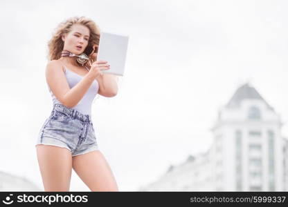 Happy young woman with vintage music headphones around her neck, taking selfie with tablet pc and smiling happily against urban city background.