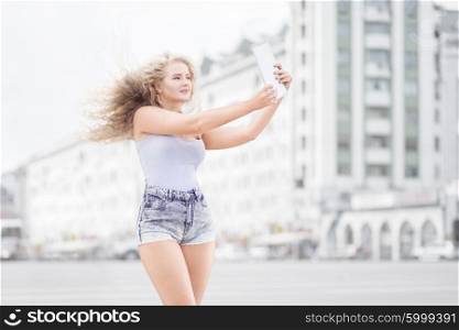 Happy young woman with vintage music headphones around her neck, taking selfie with tablet pc and smiling happily against urban city background.