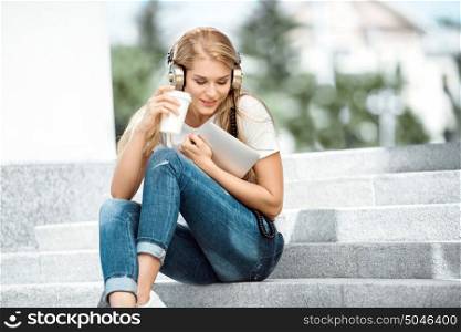 Happy young woman with vintage music headphones, holding a take away coffee cup and tablet pc, sitting on stairs against urban city background.