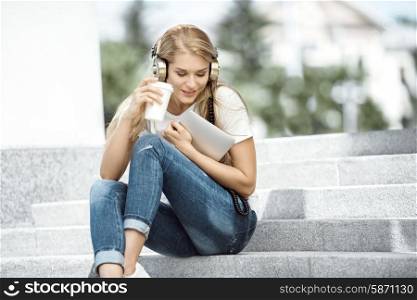 Happy young woman with vintage music headphones, holding a take away coffee cup and tablet pc, sitting on stairs against urban city background.