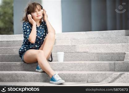 Happy young woman with vintage music headphones, listening to the music and sitting on stairs with a take away coffee cup against urban city background.
