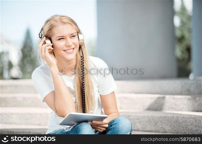 Happy young woman with vintage music headphones, surfing internet on a tablet pc and sitting on stairs against urban city background.