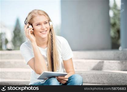 Happy young woman with vintage music headphones, surfing internet on a tablet pc and sitting on stairs against urban city background.