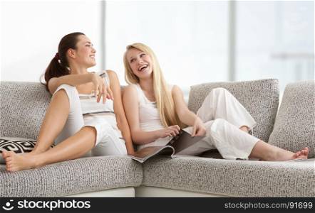 Happy Young Women Reading Fasion Magazine, Chatting, Lying on the Sofa in Living Room