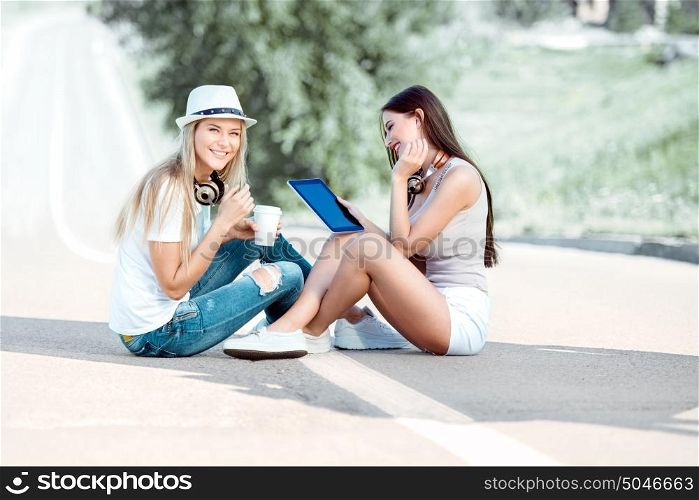 Happy young women sitting on the road, drinking coffee from a takeaway coffee cup, wearing music headphones, using tablet and buying music online against road background.