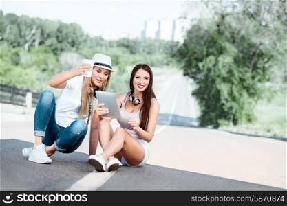 Happy young women sitting on the road, drinking coffee from a takeaway coffee cup, wearing music headphones, using tablet and buying music online against city background.