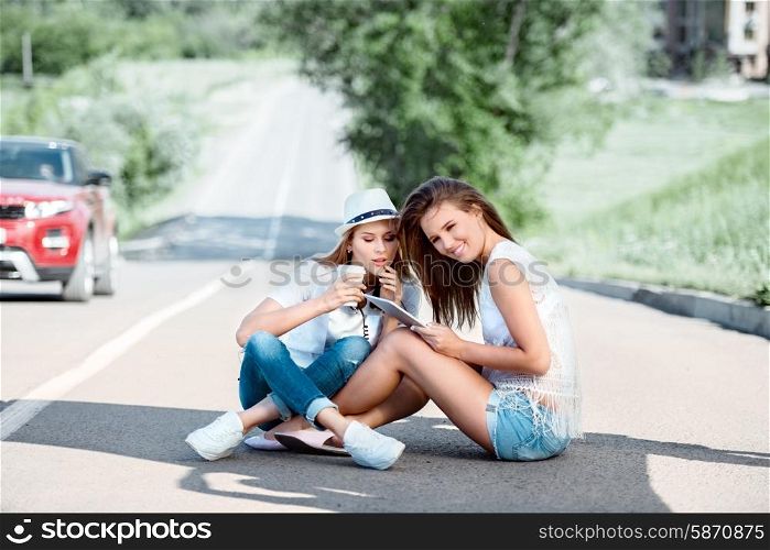 Happy young women sitting on the road, drinking coffee from a takeaway coffee cup, wearing music headphones, using tablet and buying music online against city background.