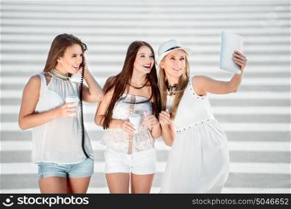 Happy young women standing on zebra crossing, drinking coffee from a takeaway coffee cup, wearing music headphones, buying music on tablet online and taking selfie against city background.