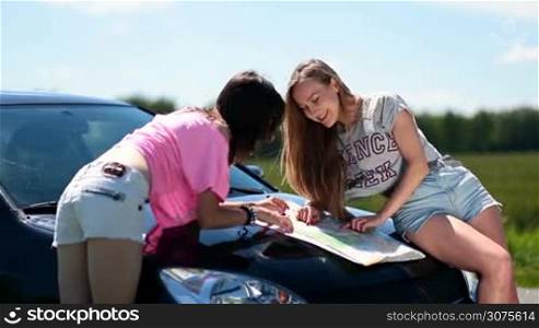 Happy young women stopped the car on the roadside of rural road and discussing their roadtrip route using a map on the engine hood of car.